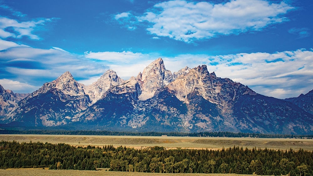 Jagged snow-capped mountain peaks rise above a forested valley under a blue sky with white clouds