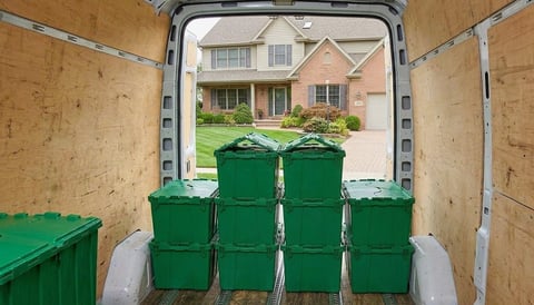 View from inside a moving truck filled with stacked green storage bins, showing a suburban brick house and driveway