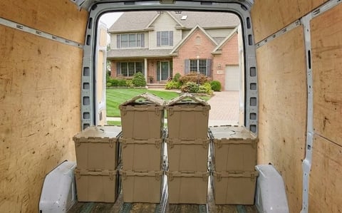 View from inside a moving truck filled with stacked green storage bins, showing a suburban brick house and driveway