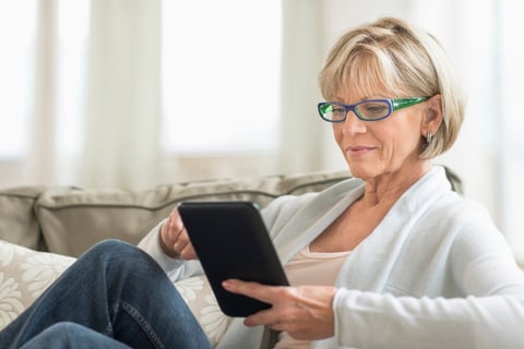 Older woman with glasses wearing white cardigan sits on couch using a tablet device